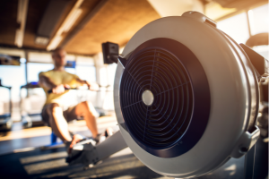 Close up focus view of rowing machine while strong motivated and focused muscular bald bodybuilder man with earphones pulling the bar in the modern sunny gym.