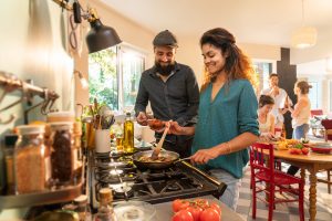 an ethnic man and woman stand over a pan on the stove top while a group of friends chat in the background