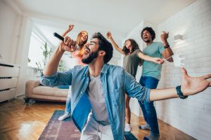 man in blue button up sings into a microphone while three friends dance behind him