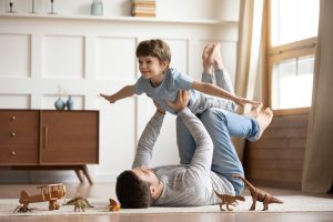 Joyful young man father lying on carpet floor, lifting excited happy little child son at home.