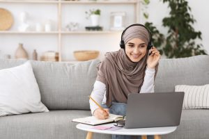 Girl studying online from her laptop wearing headphones and writing in a notepad with a computer.