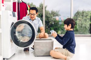 Family asian father and kid little boy son having fun doing household chores doing laundry dirty clothes into the washing machine together in laundry room at home.