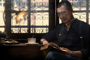 Older Asian man reads book and drinks coffee