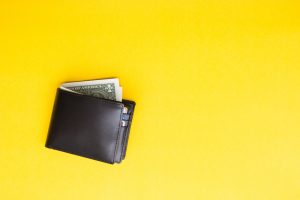 Black leather wallet with dollar cash peaking out the top on yellow background.