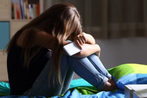 Single sad teen holding a mobile phone lamenting sitting on the bed in her bedroom with a dark light in the background representing teen anxiety.