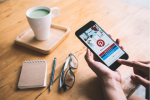 someone opens Pinterest on their phone against a desk backdrop of coffee, a notebook, pen, and pair of glasses